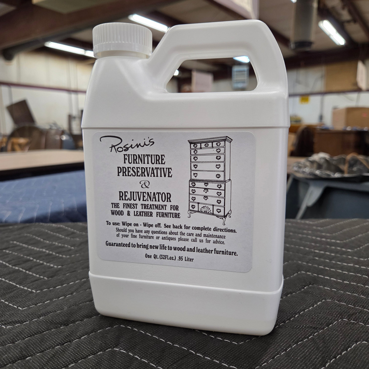 A white plastic bottle of Rosini's Furniture Preservative and Rejuvenator on a dark gray surface in a warehouse workshop.