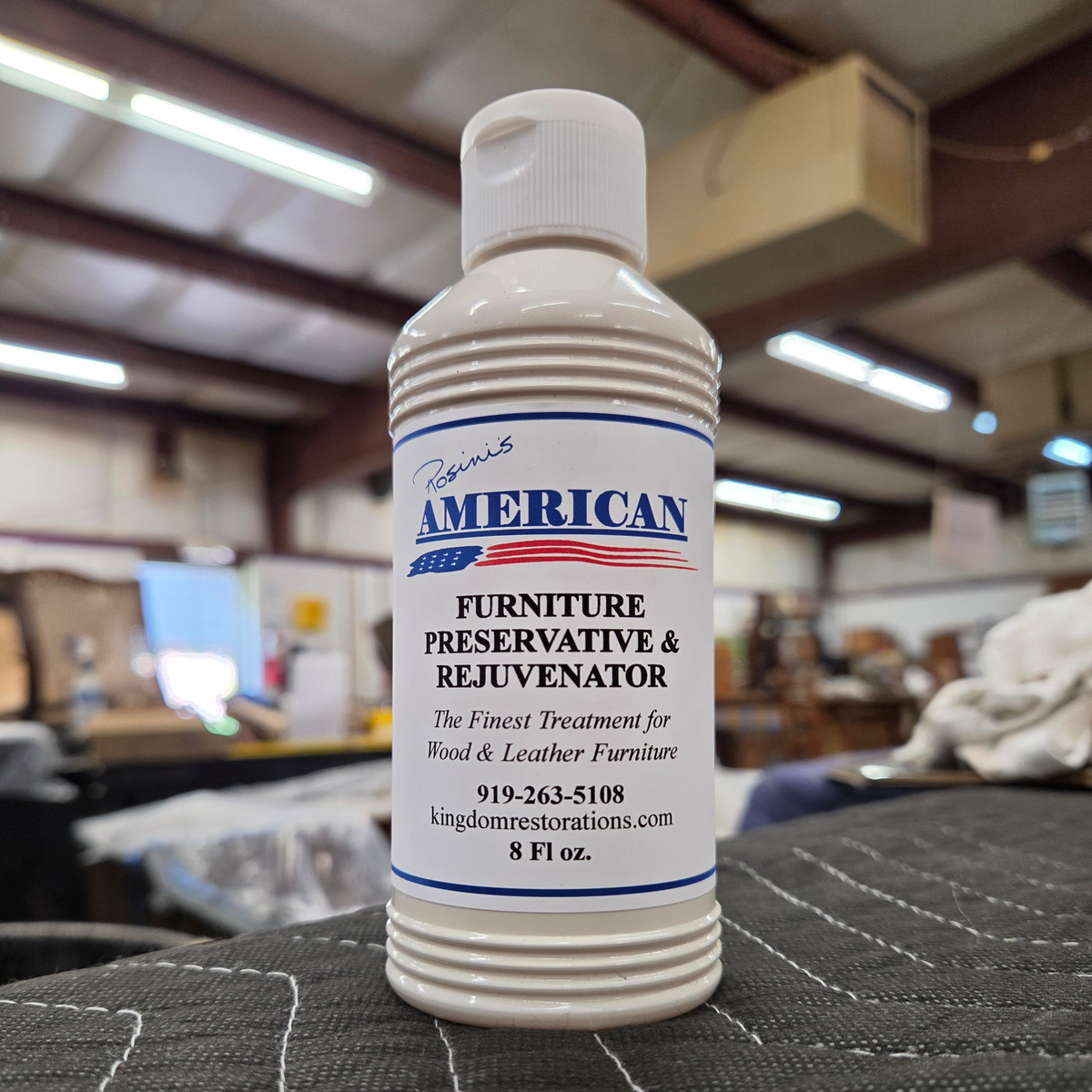 An 8 fluid ounce bottle of Rosini's American Furniture Preservative and Rejuvenator on a dark gray surface with a blurred warehouse workshop in the background.