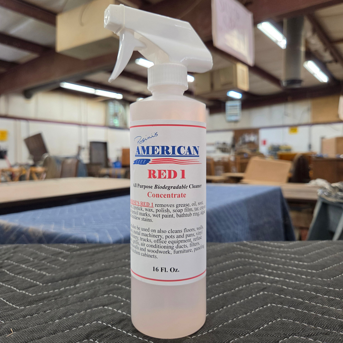 A spray bottle of Rosini's American Red 1 Concentrate, shown resting on a dark gray surface in a warehouse workshop setting.